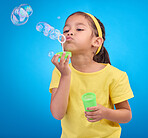 Children, blue background and a girl blowing bubbles in studio for fun or child development of motor skills. Kids, game or soap and a cute or adorable female child playing with a bubble wand 