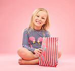Popcorn, food and happy girl portrait in a studio with pink background sitting with movie snacks. Snack, happiness and hungry child with a paper bag and chips eating and feeling relax with a smile