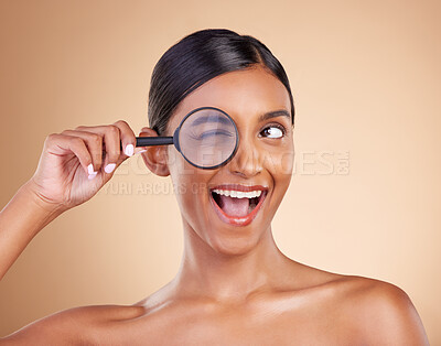 Buy stock photo Face, beauty or magnifying glass with a model woman in studio on beige background searching for skincare. Facial, eye wink or excited Indian girl looking for luxury self care cosmetics or makeup
