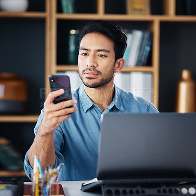 Buy stock photo Serious asian man, phone and laptop for social media, communication or networking at office desk. Male creative designer looking at smartphone by computer for browsing, research or planning startup