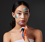 Beauty, portrait and woman with paint art on hand and face in studio. Creative skin and rainbow makeup on serious female aesthetic model on gray background for lgbtq color inspiration on hands