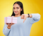 Disappointed, portrait and woman with a thumbs down for a gift isolated on a yellow background. Unhappy, negative and a girl holding a present and showing a hand gesture for disappointment in studio