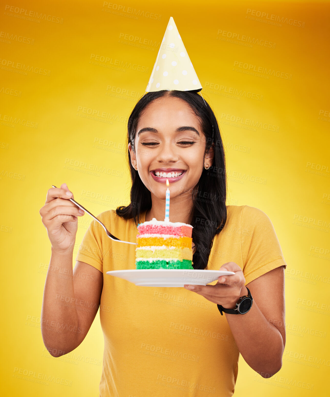 Buy stock photo Woman is eating cake, birthday celebration and happy in portrait, rainbow dessert and candle on yellow background. Celebrate, festive and young female, excited for sweet treat and party hat in studio