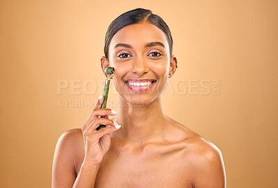 Buy stock photo Face portrait, skincare roller and woman in studio isolated on a brown background. Dermatology, facial massage and happy Indian female model with jade crystal for healthy skin treatment and beauty.