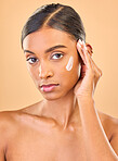 Face portrait, skincare and woman with cream in studio isolated on brown background. Dermatology, serious cosmetics and confident Indian female model with lotion, creme or moisturizer for skin health