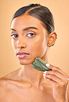 Face portrait, skincare and woman with stone in studio isolated on a brown background. Dermatology, massage and serious Indian female model with jade crystal or gua sha for healthy skin treatment.