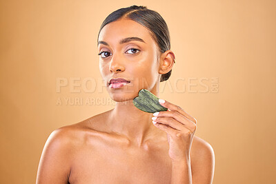 Buy stock photo Face portrait, skincare and woman with gua sha in studio isolated on a brown background. Dermatology, massage and serious Indian female model with jade crystal or stone for healthy skin treatment.