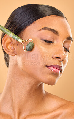 Buy stock photo Face, skincare roller and woman with eyes closed in studio isolated on a brown background. Dermatology, facial massage and Indian female model with jade crystal for healthy skin treatment and beauty.