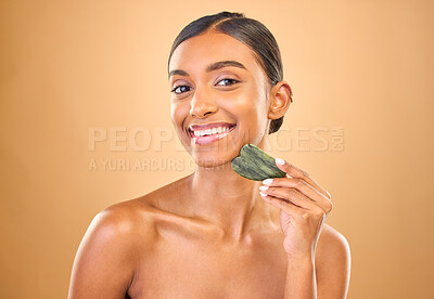 Buy stock photo Face portrait, skincare and woman with gua sha in studio isolated on a brown background. Dermatology, facial massage or happy Indian female model with jade crystal or stone for healthy skin treatment