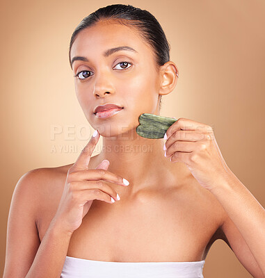 Buy stock photo Skincare, gua sha and portrait of Indian woman for beauty, facial treatment and spa wellness in studio. Salon, dermatology and serious girl on brown background with cosmetics, face massage and stone