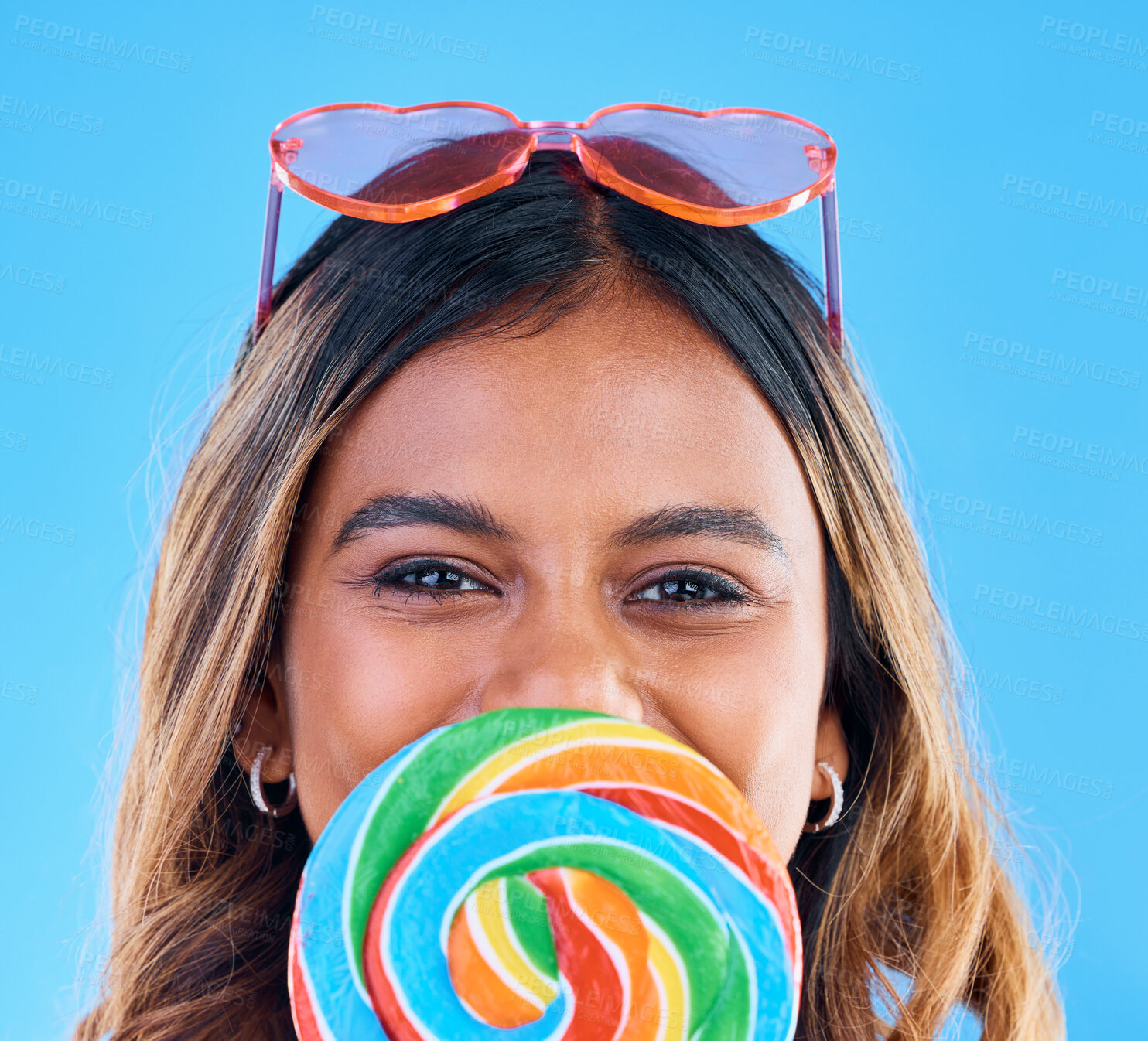 Buy stock photo Portrait, giant lollipop and a woman on a blue background in studio wearing heart glasses for fashion. Face, candy and sweet with an attractive young female eating a snack while feeling happy