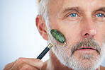 Senior man, jade roller and face for skin with smile, self care and wellness in studio by white background. Elderly model, facial massage and beauty glow with stone product, skincare and dermatology