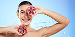 Pomegranate, portrait and water splash of woman, beauty and wellness on studio blue background. Happy model, shower and healthy fruits for natural benefits, detox nutrition and skincare dermatology