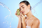 Woman, water splash and face with kiwi in relax for skincare, hygiene or nutrition against blue studio background. Calm or relaxed female holding fruit in natural beauty cosmetics or facial treatment