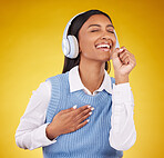 Headphones, happy and woman singing in studio while listening to music, playlist or album. Happiness, smile and Indian female model doing karaoke while streaming a song isolated by yellow background.