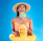 Pool float, happy woman and lollipop lick in a studio with sweet and swimsuit with a smile. Isolated, blue background and holiday outfit of a young female with happiness and candy feeling fun