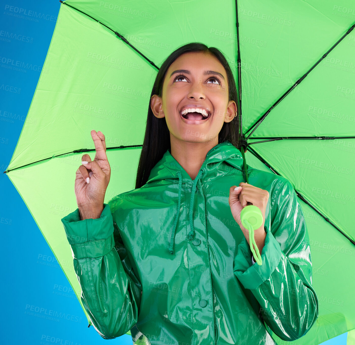 Buy stock photo Green, umbrella and fingers crossed with a woman on a blue background in studio during winter for insurance. Rain, fashion or cover and an attractive young female wishing for weather change with hope