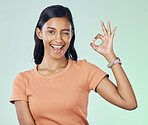 Portrait, wink and female with a perfect gesture in studio with a positive, confident and happy mindset. Happiness, smile and young Indian woman model with a ok hand sign isolated by green background