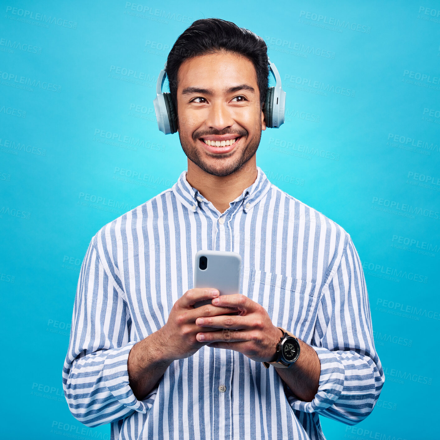 Buy stock photo Headphones, music and thinking man with phone in studio for streaming, audio and subscription against blue background. Radio, smile and happy asian male online for podcast, track and internet app