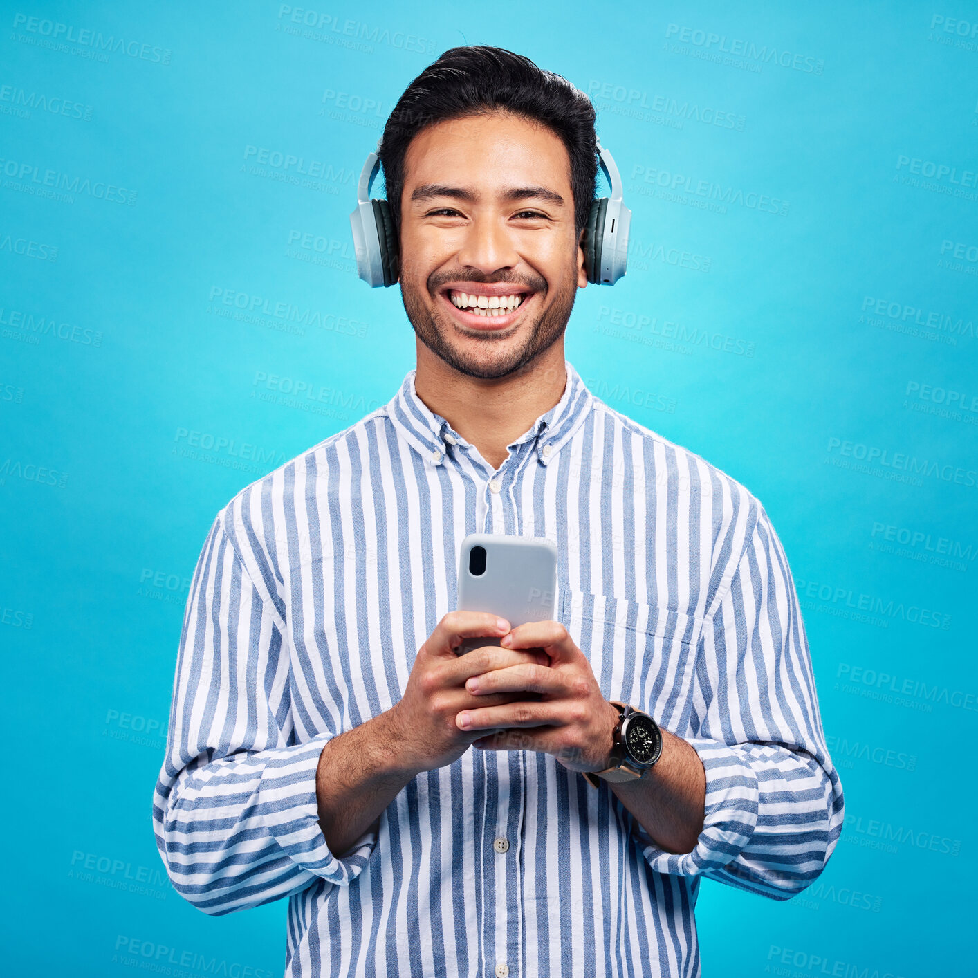 Buy stock photo Music, headphones and happy man with phone in studio for streaming, audio and subscription against blue background. Radio, smile and face of asian male online for podcast, track and internet app