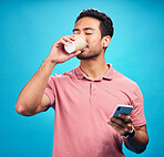 Earphones, drinking coffee and man with phone in studio isolated on a blue background. Tea, cellphone and Asian person drink, caffeine or espresso with mobile for social media, radio music or podcast