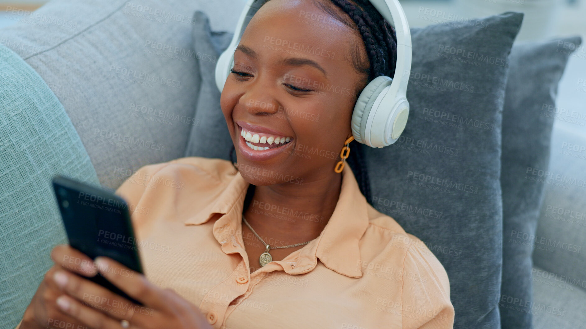 Buy stock photo Happy black woman, headphones and relax with phone on sofa for funny joke in living room at home. African female person smile on mobile app and laughing for social media, meme or entertainment
