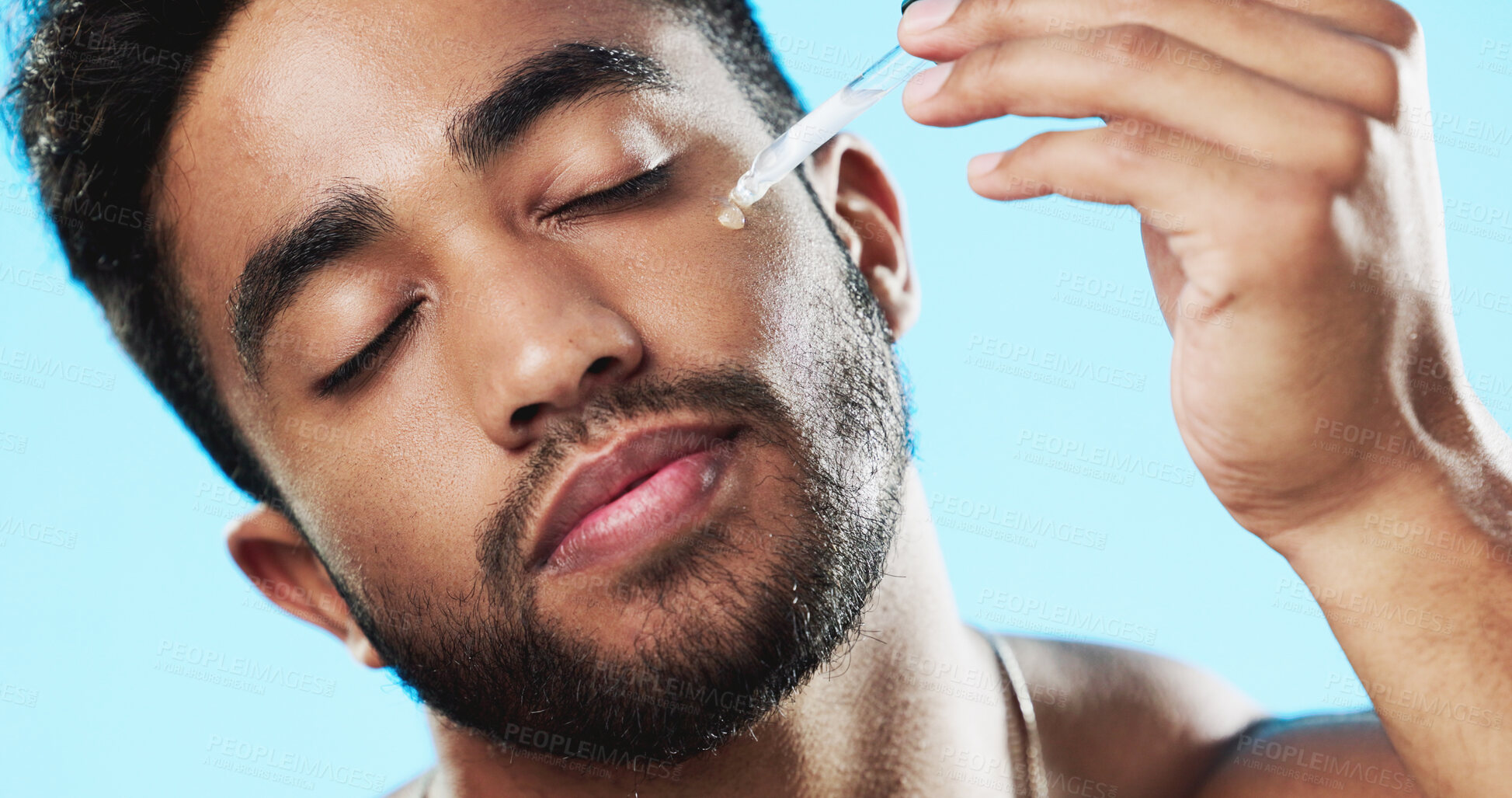 Buy stock photo Man, beauty and oil on face in studio for aesthetic skincare, dermatology and healthy glow on blue background. Indian model, facial cosmetics and dropper of hyaluronic acid, liquid serum and collagen