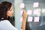 Business woman, writing and planning for brainstorming, strategy or ideas on glass board at the office. Female employee working on tasks for reminder, sticky note or project plan at the workplace
