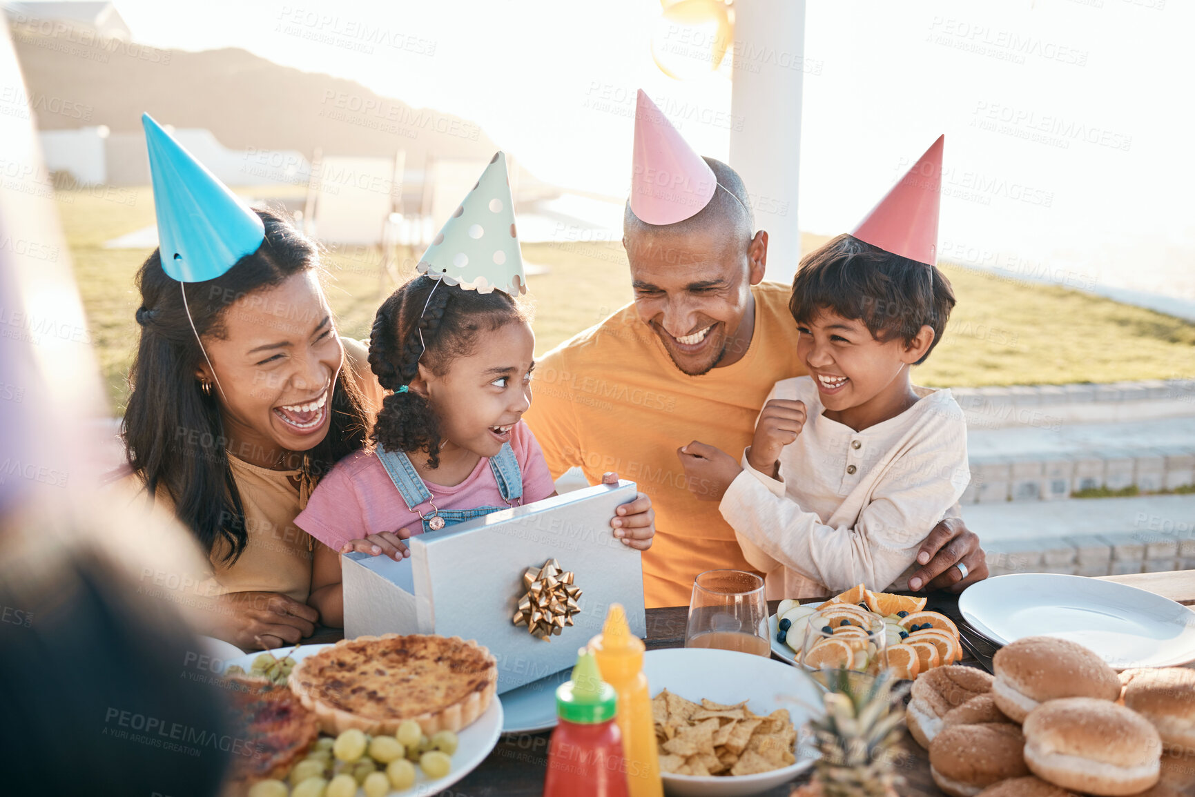 Buy stock photo Happy birthday, love and family outdoors for celebration, party and present, laugh and excited. Parents, children and gift for girl child at special event, smile and celebrating with food outside