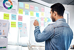 Creative man, sticky note and glass board for schedule tasks, planning or brainstorming at the office. Male person or employee checking plan, agenda or marketing strategy for startup at the workplace