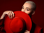 Portrait, fashion and hat with a woman on a red background in studio for elegant or trendy style. Face, hide and cover  with a young female model looking edgy or classy in a suit or unique clothes
