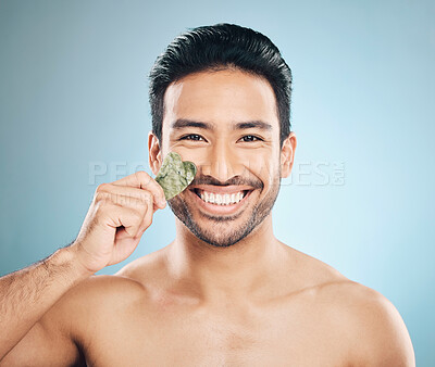 Buy stock photo Face, man and beauty, gua sha and cosmetic tools, smile in portrait and natural treatment on blue background. Skincare, healthy glow and jade stone, dermatology and wellness with male model in studio