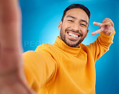 Buy stock photo Selfie, happy and peace sign portrait of a man in studio with hand, emoji and a smile. Male asian fashion model on a blue background with a positive mindset for social media profile picture update