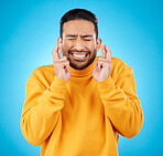 Nervous, eyes closed and fingers crossed by asian man with anxiety in studio for news, feedback or review on blue background. Hand, emoji and guy with stress for results, competition or giveaway