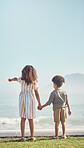 Back, holding hands and a brother with his sister on the beach, looking at a view of waves together in summer. Kids, love and pointing with a girl showing a boy the horizon on a blue sky background