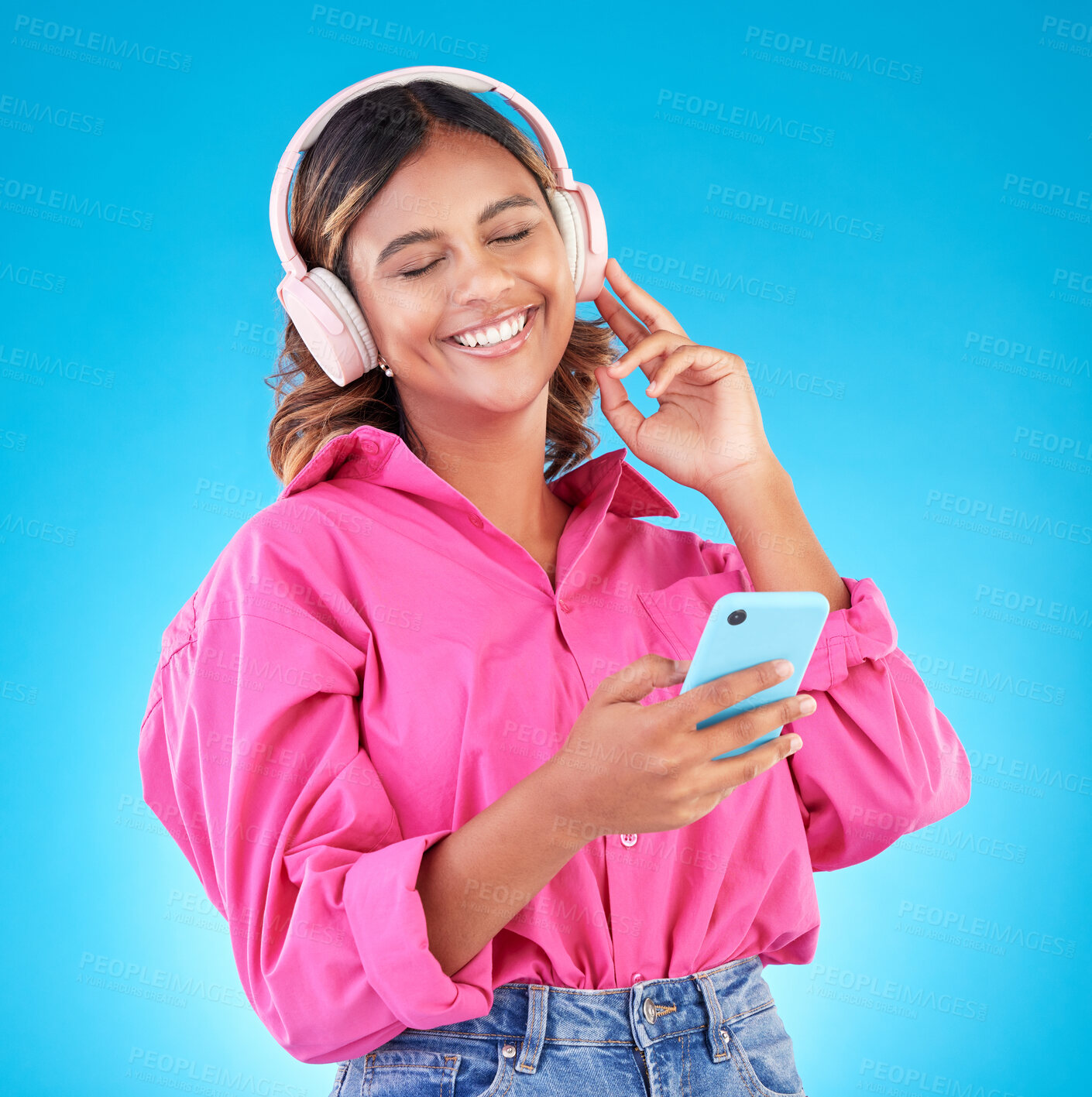 Buy stock photo Phone, music and headphones with a woman in studio on a blue background, listening to the radio. Mobile, smile and a happy young female person streaming audio to relax for wellness or freedom