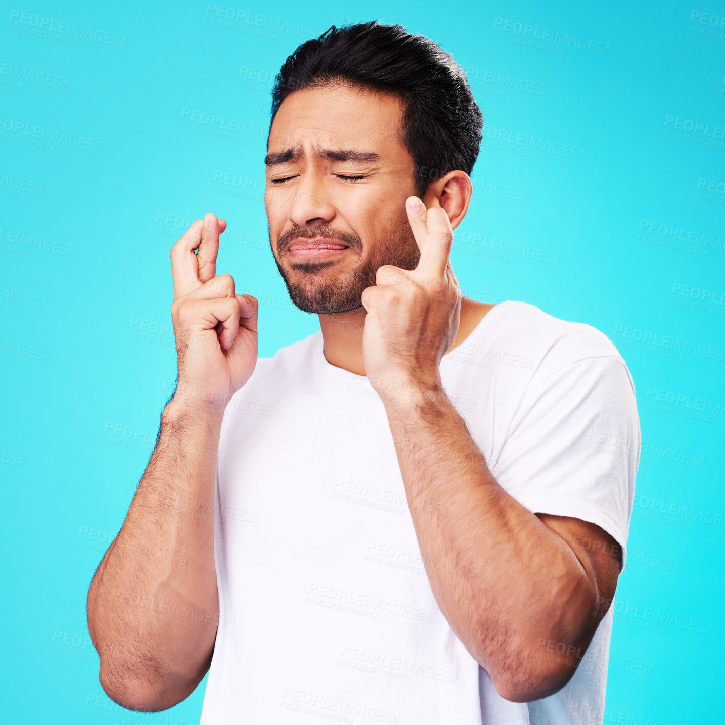 Buy stock photo Praying, hope and man fingers crossed, anxiety or nervous due to crisis or stress isolated in studio blue background. Fear, suspense and young male person hands in face for mistake or risk reaction