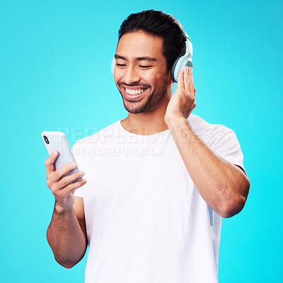 Buy stock photo Headphones, music and Asian man with smartphone for listening in studio isolated on a blue background. Radio, smile and person with mobile, sound or hearing podcast, audio or social media on internet
