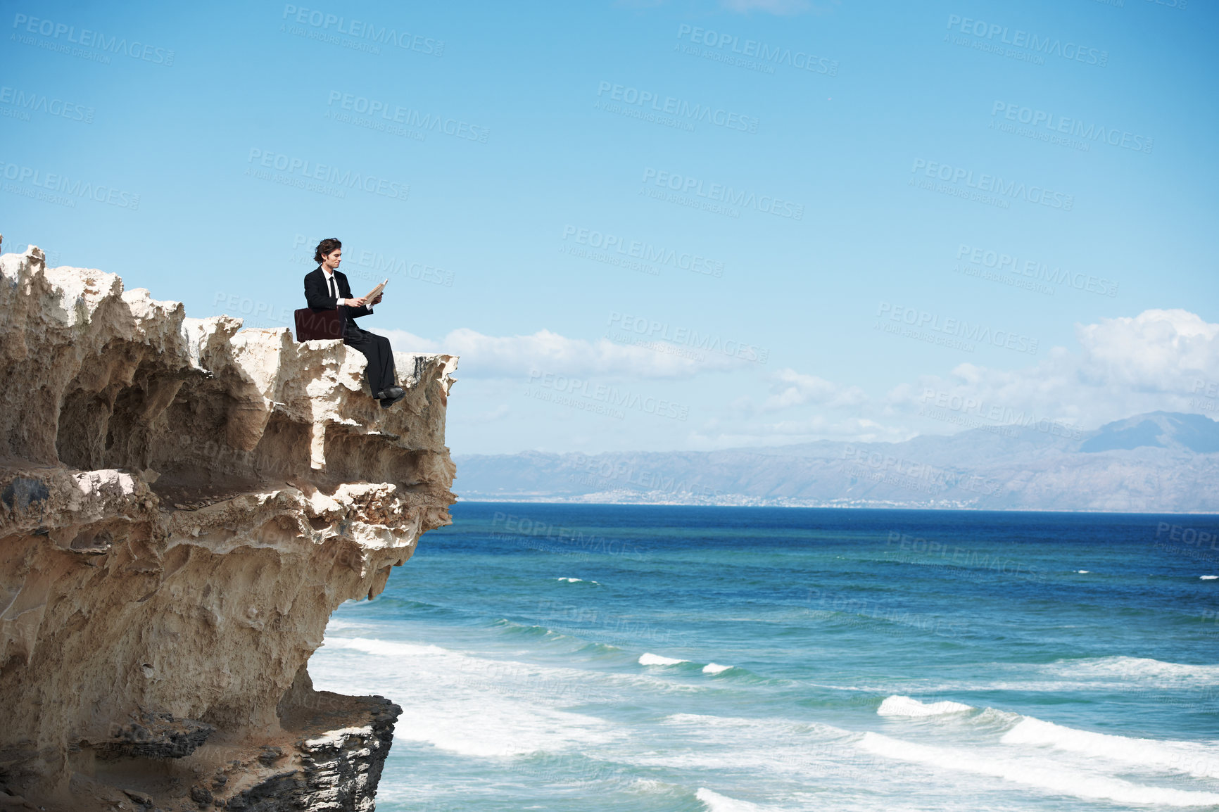 Buy stock photo Young businessman sitting on the edge of a cliff overlooking the ocean while reading a book