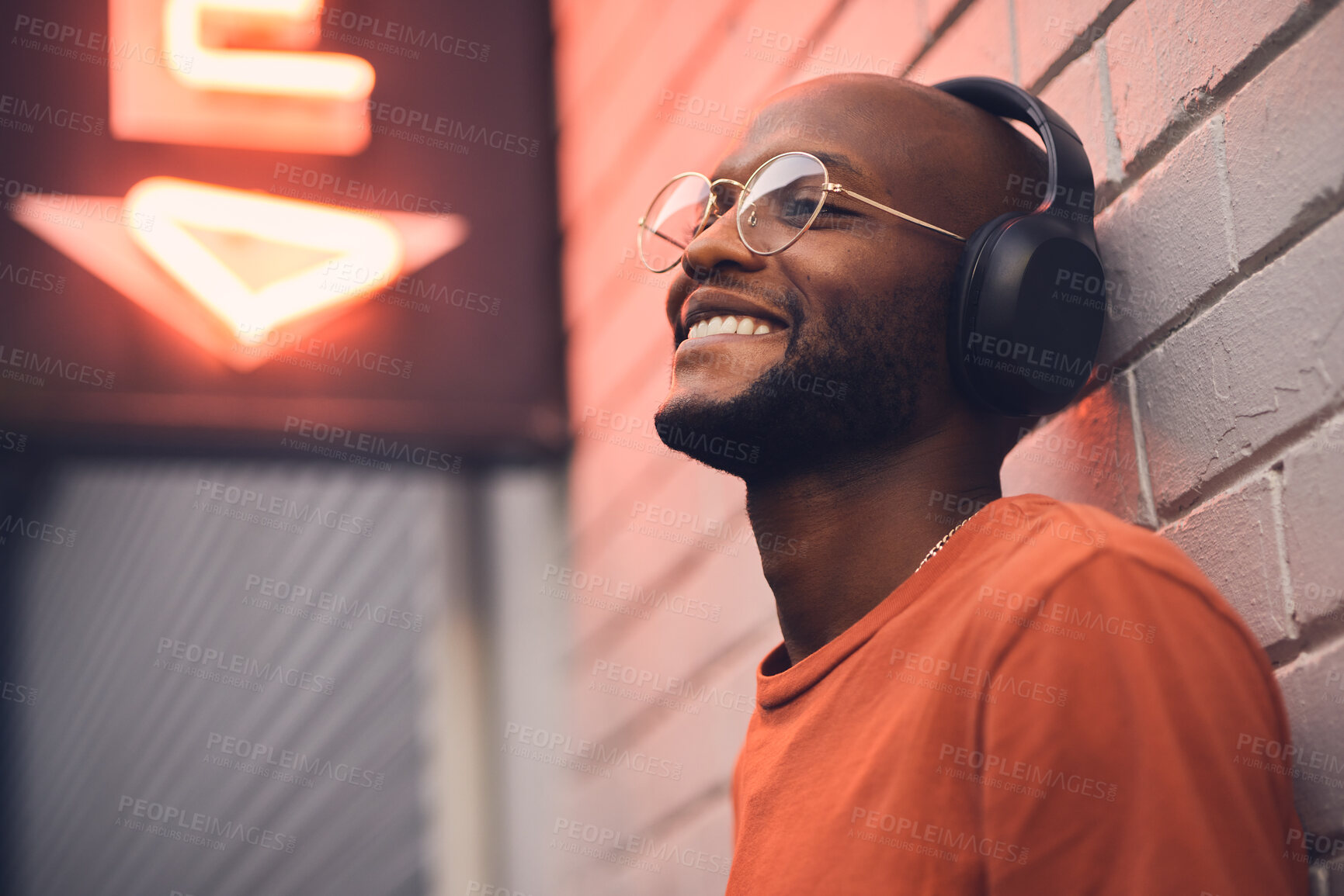 Buy stock photo Thinking, smile and music with a black man in the city, leaning on a brick wall on the street at night. Idea, glasses and headphones with a happy young male person streaming or listening to audio