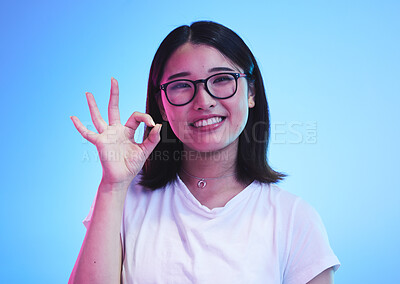 Buy stock photo Woman, okay hands or sign for education success, certified and excellence in tshirt and a blue, studio background. Happy young, asian person in glasses, portrait and yes or support emoji for learning