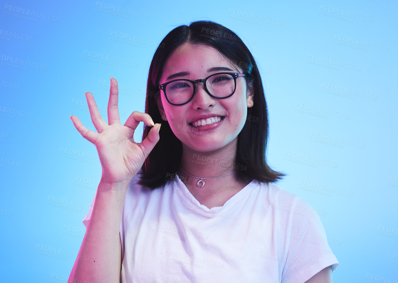 Buy stock photo Woman, okay hands or sign for education success, certified and excellence in tshirt and a blue, studio background. Happy young, asian person in glasses, portrait and yes or support emoji for learning