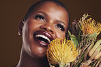 Beauty, makeup and black woman with flowers in a studio for natural face cosmetics for self care. Happy, smile and young African female model with floral aesthetic isolated by a brown background.
