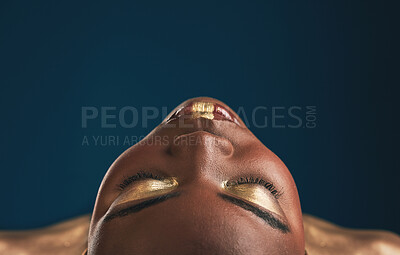 Buy stock photo Beauty, gold makeup and art on face of black woman with creative gold aesthetics isolated in a studio blue background. Artistic, cosmetic and young person eyes closed for fantasy with color or paint