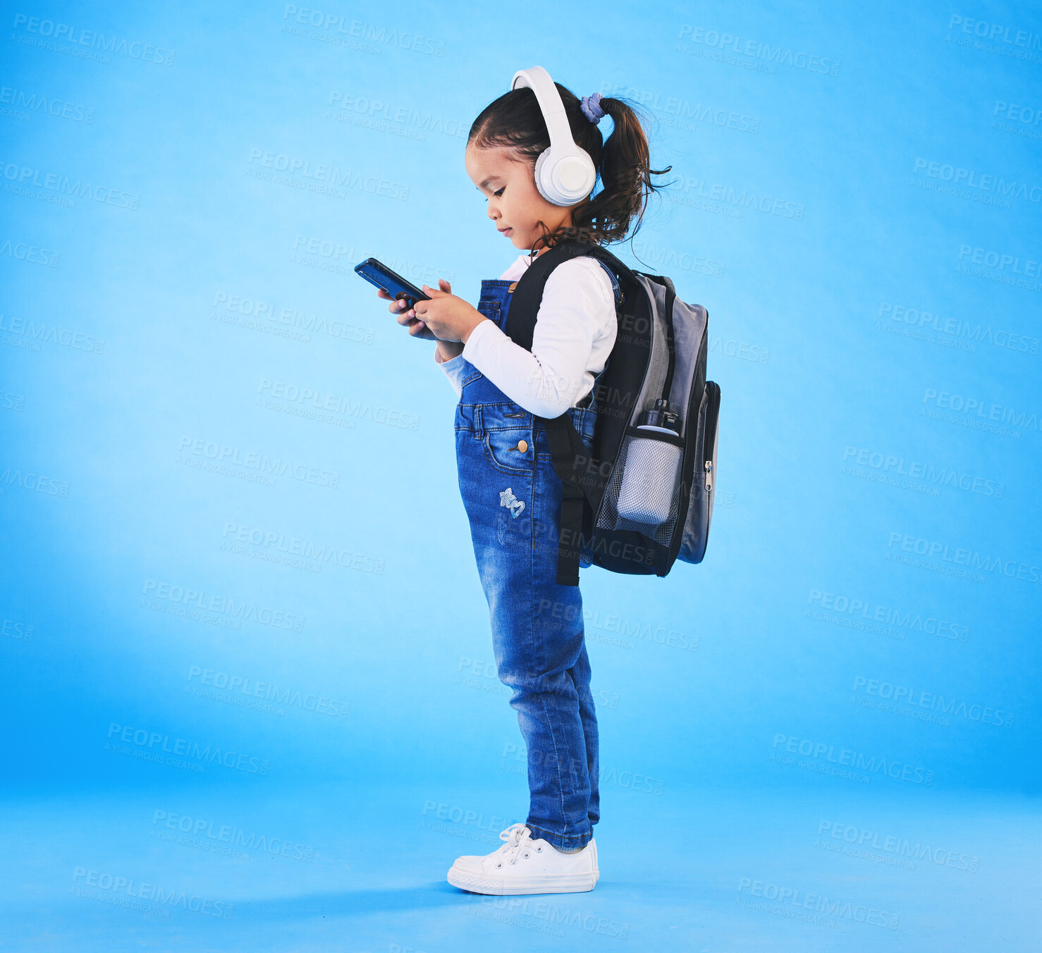 Buy stock photo Headphones, backpack and child on blue background with phone ready for school, learning and education. Kindergarten, and young girl with bag on smartphone for streaming music, audio and radio