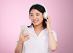 Headphones, smile and woman with a phone in a studio listening to music, playlist or album for entertainment. Happy, calm and Asian female model streaming a song or radio isolated by pink background.