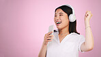 Headphones, singing and woman with a phone in a studio listening to music, playlist or radio. Happy, smile and young Asian female model doing karaoke with a cellphone mic to a song by pink background