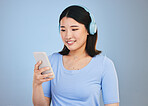 Woman, phone and headphones for listening, audio streaming and social media on a blue background. Young asian person with music subscription, electronics and mobile chat or web networking in studio