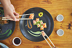 Chopsticks, hand and eating sushi and food at a restaurant for nutrition at table. Above hungry people with wooden sticks and soy sauce for dining, Japanese culture and cuisine with art on a plate