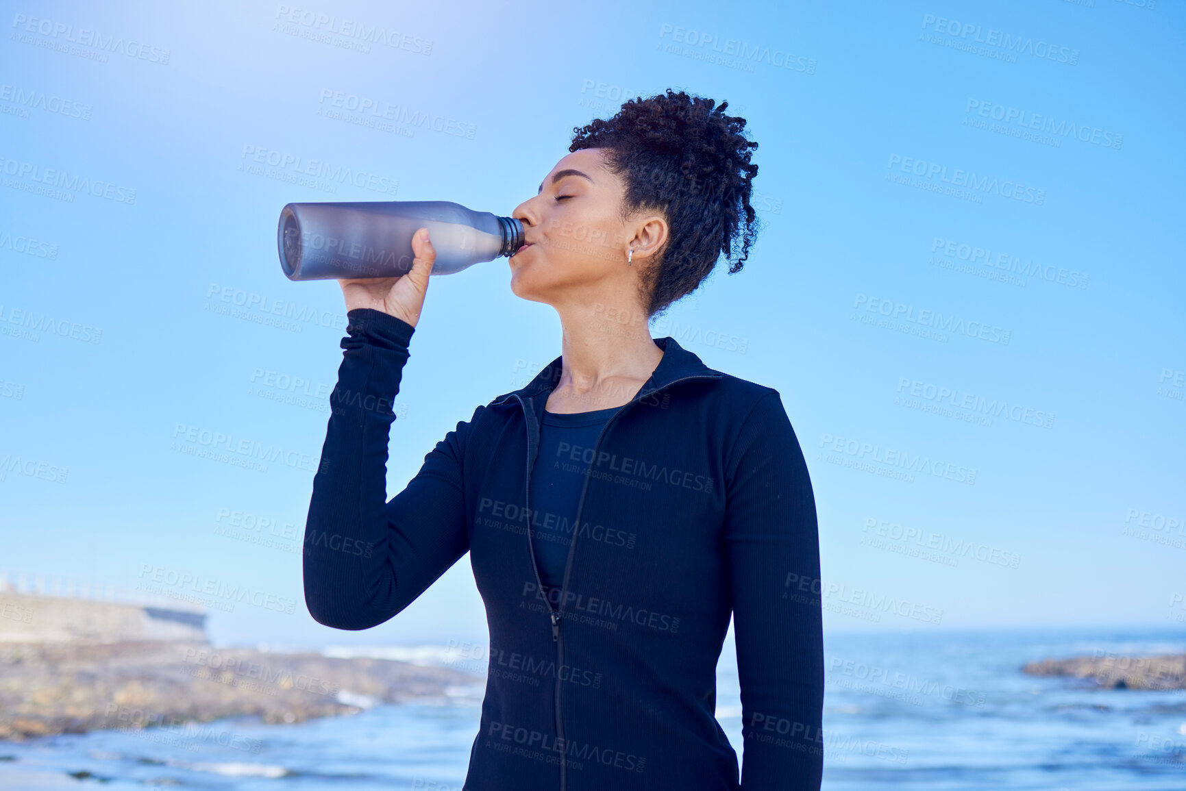 Buy stock photo Fitness, woman and drinking water at beach for running, health or morning cardio on blue sky by an ocean. Exercise, hydration and thirsty lady runner with sports liquid after training or sea workout